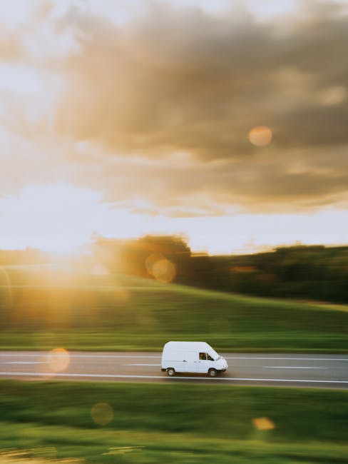 A white commercial van operated by Man with Van Edmonton is seen in motion on a multi-lane road, with a blurred background of a brick wall and street features indicative of an urban environment. The van is positioned centrally in the image, with its side doors visible, suggesting a home relocation or furniture transport process. The vehicle is equipped for removals services, suitable for loading household furniture, boxes, and packaging materials. The setting appears to be daytime, with natural lighting illuminating the vehicle, and the blurred motion implies high speed typical of fast removals or logistics operations. This image captures the essence of professional removals, emphasizing efficient vehicle use in the urban moving industry, aligning with the topics covered in Edmonton Green: Best Van Routes for Fast Removals, EDMONTON from [COMPANY_NAME].