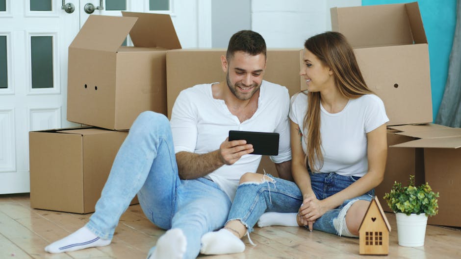 A young couple with light skin sitting on a light wooden floor inside a home, surrounded by open cardboard boxes used for house packing and moving. The man, wearing a white t-shirt and light blue jeans, is holding a smartphone and smiling, while the woman, dressed in a white top and ripped jeans, sits close beside him, looking at the device. Behind them are several large cardboard boxes in various states of being packed, some open and empty, with others closed, indicating a home relocation process. To the right, there is a small plant in a white pot and a miniature wooden house model, both placed on the floor. The scene captures the interior of a residential space with a white door and interior wall visible in the background, highlighting the packing stage of furniture transport and house removals facilitated by Man with Van Edmonton, demonstrating the packing and loading process involved in professional removals services.