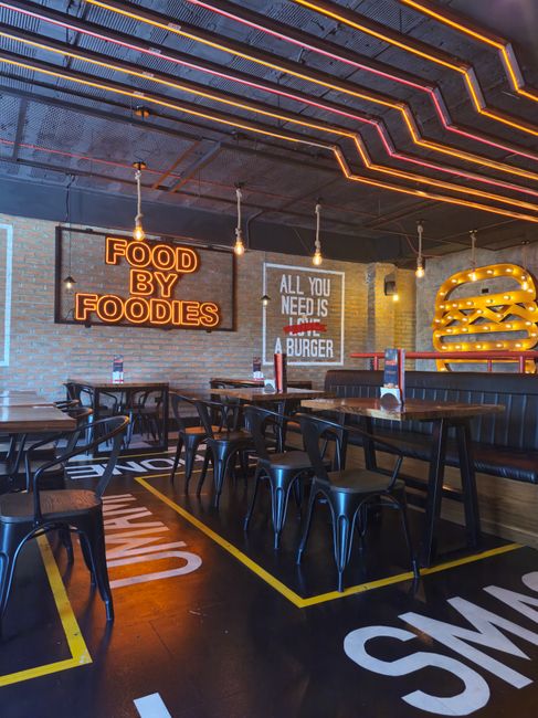 Interior of a casual dining restaurant with exposed brick walls and colorful lighting on the ceiling. Several dark wooden tables and black metal chairs are arranged on a black floor marked with yellow lines indicating seating zones. Neon signs on the walls include one saying 'FOOD BY FOODIES' in bright orange, and another sign mentioning 'ALL YOU NEED IS A BURGER' in white and red lettering. A large decorative burger sculpture made of illuminated yellow and black material is mounted on the wall. The environment appears ready for a home relocation or moving services involving packing and transport, with visible lighting and industrial decor elements consistent with a modern, urban eatery.