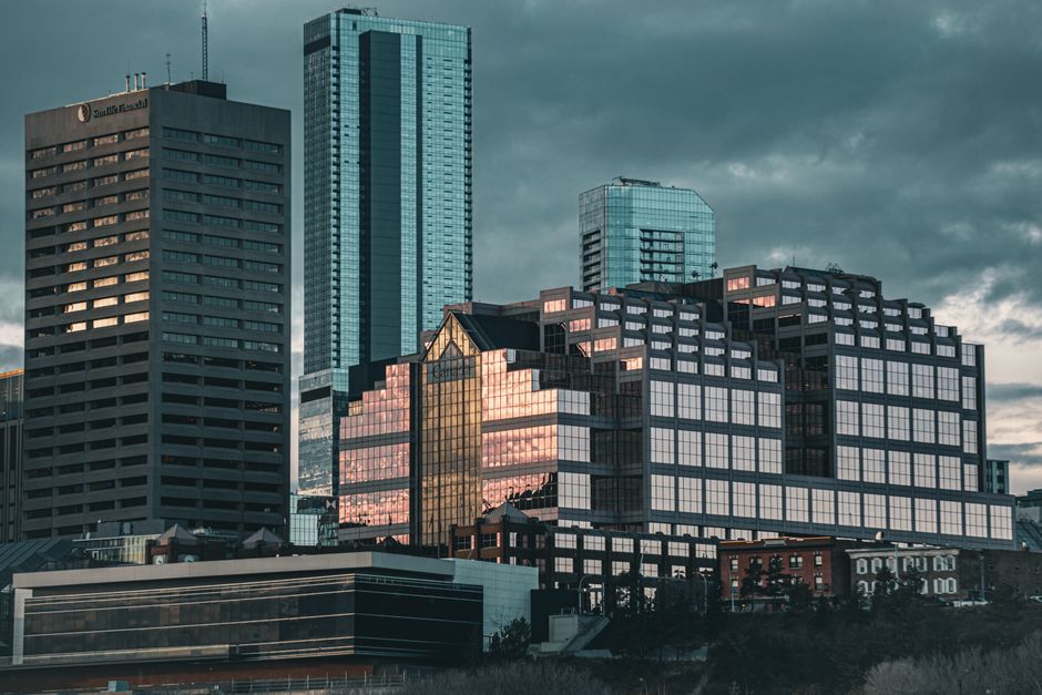 A cityscape featuring multiple modern high-rise buildings with glass and concrete facades, some reflecting the setting sun's warm light. The lower part of the image shows a park with leafless trees an