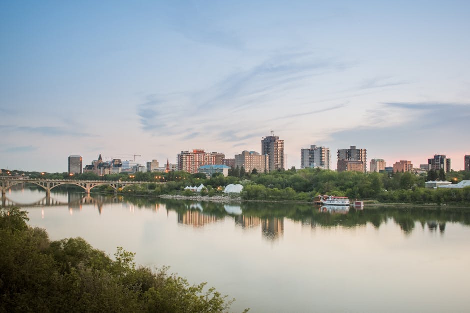 A panoramic view of a city skyline across a river during the early evening, with several high-rise buildings, some under construction with cranes visible, and a variety of modern office and residential towers. The river in the foreground reflects the cityscape, with a calm water surface and green foliage along the riverbank. A bridge with arches spans across the river on the left side, and a small boat is moored near the grassy shoreline. Inside a property, there are cardboard boxes, wrapped furniture, and moving blankets arranged near an open doorway, with a hand truck positioned nearby as part of a home furniture transport or packing and moving process. The scene is lit by natural light, indicative of the late sunset or dusk, and the environment suggests a house removal or relocation activity taking place, aligned with professional removals services such as those offered by Man with Van Edmonton.