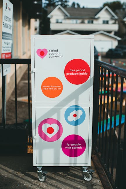A white, two-door public mailbox with colorful graphic signage attached, situated outdoors on a paved area near metal fencing. The signage includes a large pink heart symbol and text promoting a period pop-up event in Edmonton, with phrases such as 'free period products inside!', 'take what you need, leave what you can', and 'for people with periods.' The mailbox is mounted on four small caster wheels, suggesting it is mobile or portable for placement during moving or community events. In the background, there is a residential area with houses and cars parked along the street, and the scene is lit by natural daylight, indicating daytime. This image is relevant to house removals, highlighting the importance of accessible community facilities during relocation processes, and demonstrates practical and inclusive signage typically used in packing and moving logistics by a company like Man with Van Edmonton.