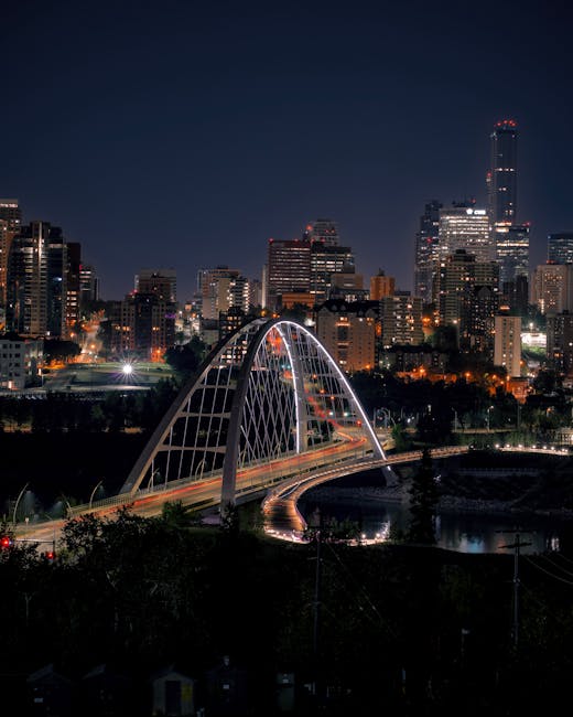 A panoramic view of a city skyline across a river during the early evening, with several high-rise buildings, some under construction with cranes visible, and a variety of modern office and residential towers. The river in the foreground reflects the cityscape, with a calm water surface and green foliage along the riverbank. A bridge with arches spans across the river on the left side, and a small boat is moored near the grassy shoreline. Inside a property, there are cardboard boxes, wrapped furniture, and moving blankets arranged near an open doorway, with a hand truck positioned nearby as part of a home furniture transport or packing and moving process. The scene is lit by natural light, indicative of the late sunset or dusk, and the environment suggests a house removal or relocation activity taking place, aligned with professional removals services such as those offered by Man with Van Edmonton.