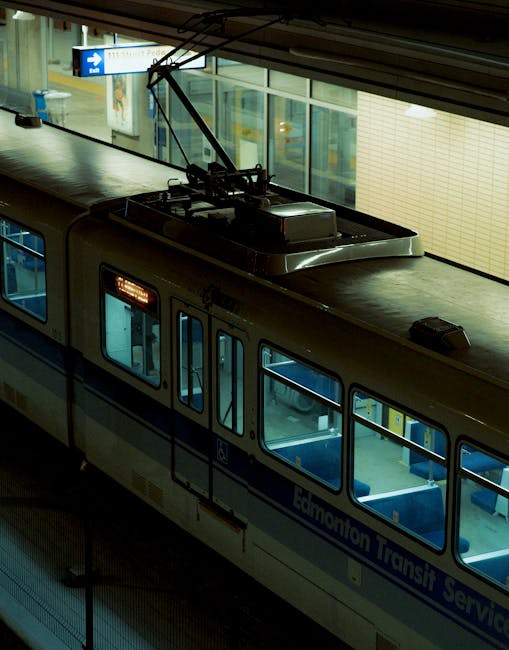 A close-up view of a light grey Edmonton Transit Service train carriage parked at an indoor platform, with large windows revealing the interior seating and lighting. The train's roof features a pantograph connecting to overhead electric lines, used for powering the vehicle. In the background, glass windows and signage are visible through the station's interior, indicating the station environment. The scene is illuminated with artificial lighting, emphasizing the clean and modern appearance of the transportation vehicle, which is associated with urban transit and city logistics. This image is relevant for house removals and moving services, illustrating the transportation methods involved in furniture and household items relocation, as provided by companies such as Man with Van Edmonton.