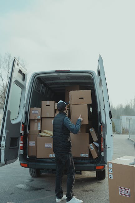 A man wearing a dark vest, black pants, and a baseball cap is inside the rear of a white delivery van with its back doors open, visible on a paved area outside a property during a home relocation process. He appears to be reaching for or adjusting a medium-sized cardboard box with a handle cutout, which is one of several similar boxes stacked inside the van. The van's cargo space contains multiple cardboard boxes of various sizes, some sealed with packing tape and labeled with barcodes and fragile stickers. A few boxes are positioned near the van's floor, while others are stacked towards the ceiling, indicating an active loading or unloading process. Outside the vehicle, the environment is overcast, with a cloudy sky and a background of trees and a distant building, suggesting an outdoor setting typical of house removals. The image subtly illustrates furniture transport and packing activities associated with professional removal services offered by Man with Van Edmonton, aligning with the theme of same-day removals in Edmonton.
