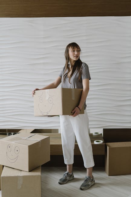 A young woman with long brown hair, dressed in a casual grey t-shirt, white cropped trousers, and grey sneakers, is standing indoors holding a medium-sized cardboard moving box with a simple smiley face drawn on it in black marker. She is positioned in front of a modern, textured white wall, and appears to be in a home environment prepared for a house relocation. On the floor nearby, there are two additional cardboard boxes, one of which also has a smiley face drawn on it, suggesting the process of packing for a move. The woman’s stance indicates she is about to load or transport the box, reflecting the furniture transport and packing activities typical of home removals. To the right, there is a dark wooden bench, and the scene is illuminated by soft, natural lighting, conveying an organized and calm moving environment. This setup aligns with professional house removals services, such as those offered by Man with Van Edmonton, specialising in same-day relocations in Edmonton.