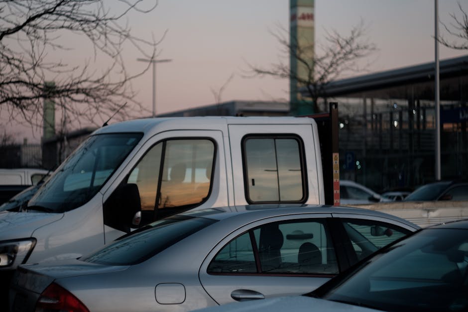 A white panel van with a sliding side door is parked in a loading area next to a silver sedan. The van's side mirror is visible, and the vehicle appears to be in the process of a home relocation, with the rear cargo area open and partially visible behind the cab. The silver car is parked in front of the van, with its windows reflecting the surrounding environment. In the background, there is a modern building with large glass windows and a covered parking area, along with a few leafless trees and a pole, suggesting it is late afternoon or early evening. The scene is lit by soft natural light, and some of the furniture or boxes typically associated with removals are not visible in this particular image, but the context aligns with vehicle loading for moving services offered by Man with Van Edmonton as part of home packing and furniture transport.
