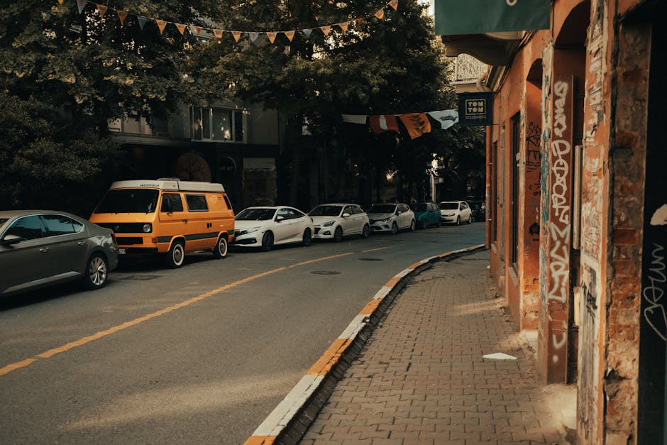 A narrow urban street scene showing a row of parked cars including a silver hatchback, a white sedan, and an orange van on a paved road with a broken yellow line. To the right, a brick building with graffiti-covered walls features a storefront with a green awning and a partially visible door. The sidewalk, made of interlocking paving stones, runs parallel to the parked vehicles, which are close to the building. Overhead, a string of decorative triangular flags is strung across the street, and trees cast shade over the area. This setting reflects a typical residential and commercial area where local house removals or furniture transport may take place, with the street available for loading and unloading activities for professional moving services like Man with Van Edmonton, who specialises in home relocation logistics and packing support.