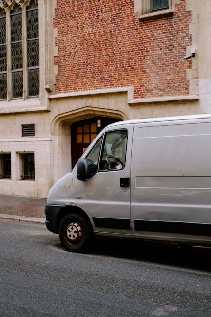A white panel van is parked on a paved street in front of a brick building with large windows and an arched doorway. The van is positioned close to the curb, with no visible signage or markings. The b
