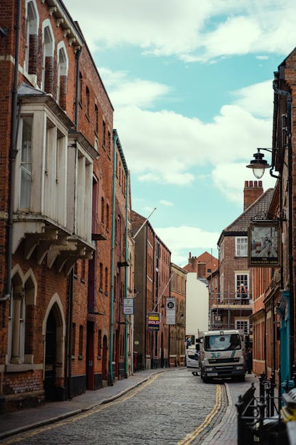 A white panel van is parked on a paved street in front of a brick building with large windows and an arched doorway. The van is positioned close to the curb, with no visible signage or markings. The b
