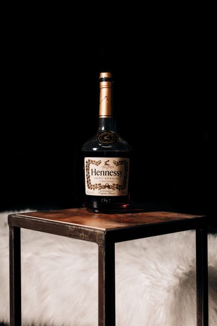 A close-up of a large Hennessy cognac bottle with a gold and black label, placed on a small wooden table with a dark metal frame. The bottle is positioned centrally on the table, which is situated on a white, fluffy surface. The background is entirely black, highlighting the bottle and table. The scene is well-lit, emphasizing the bottle's glossy surface and label details. This setup could be part of a home decor or packing process during a house move, with the table used for packing or storing items before transport. Man with Van Edmonton, a professional removals company, can assist with furniture transport and efficient packing during house relocations.